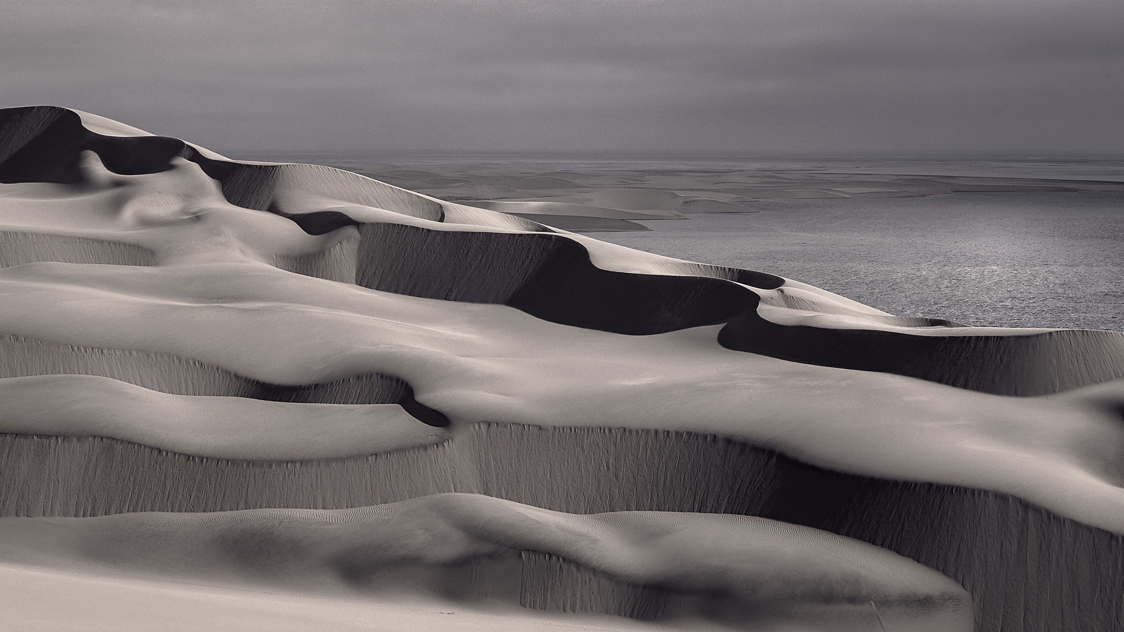 Panoramic fine art photograph of sweeping coastal sand dunes with dramatic light and shadow, overlooking the ocean under a moody sky