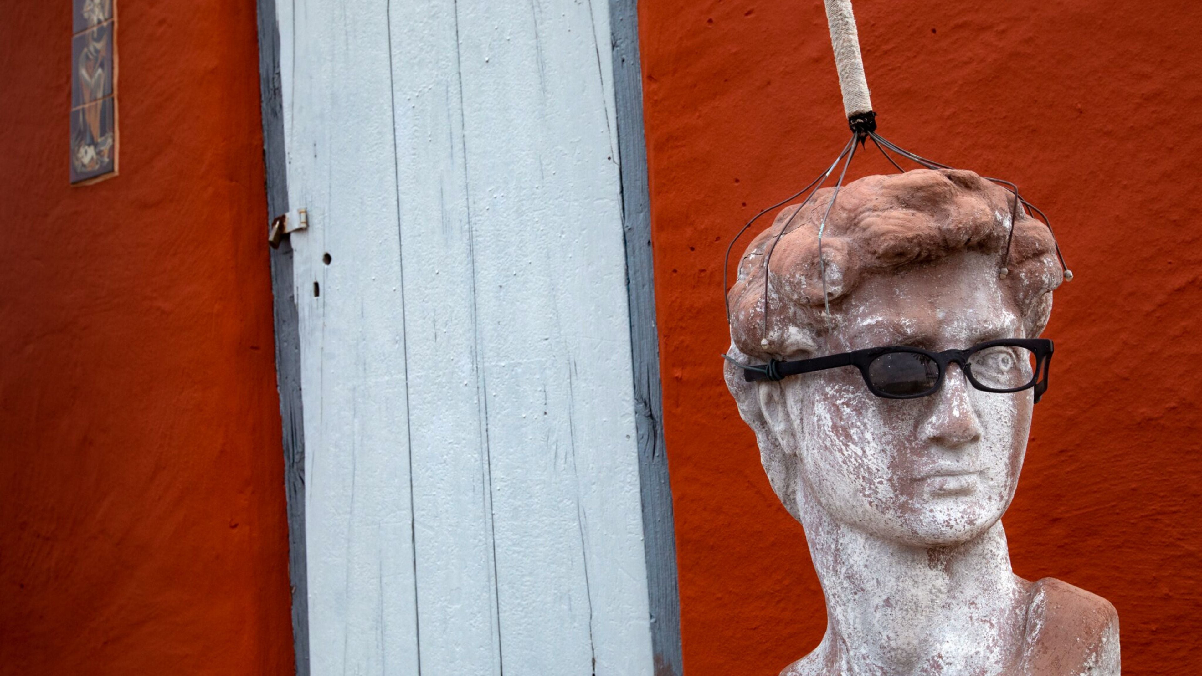 Quirky fine art photograph of a classical bust wearing black glasses, hanging against a bright red wall and pale wooden door