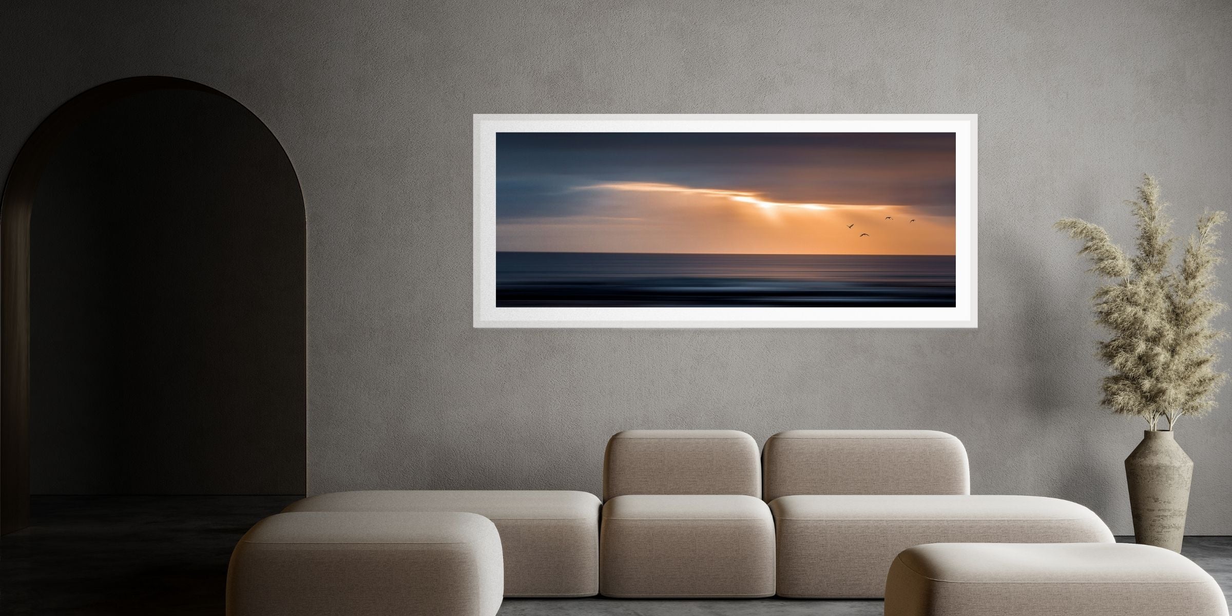 Framed artwork of a sunset over water on a gray wall above a beige sofa.